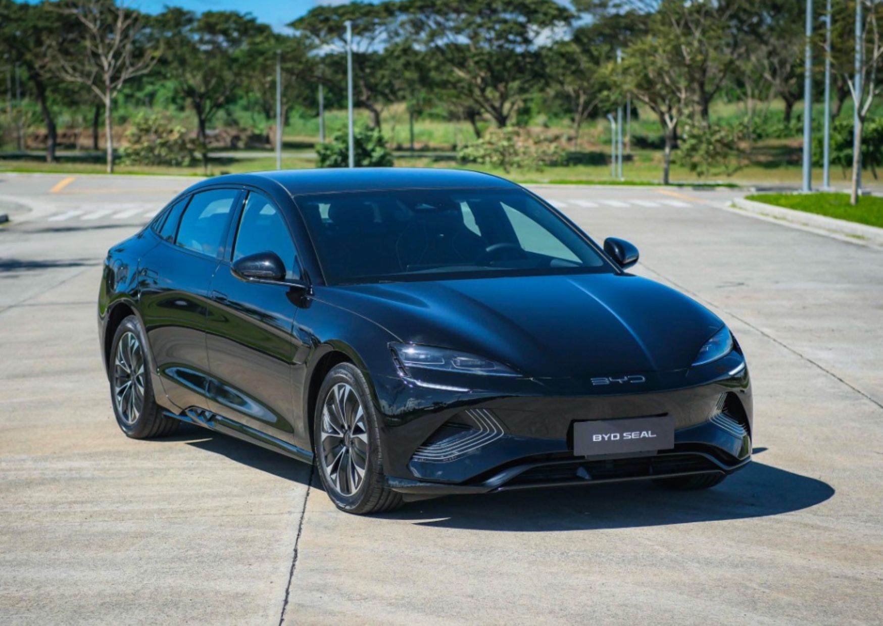 This is the BYD Seal Advanced sports sedan, an EV with a rear-wheel drive powertrains. The picture shows the car in a Picturesque background with trees and a clean pavement under a bright daylight.