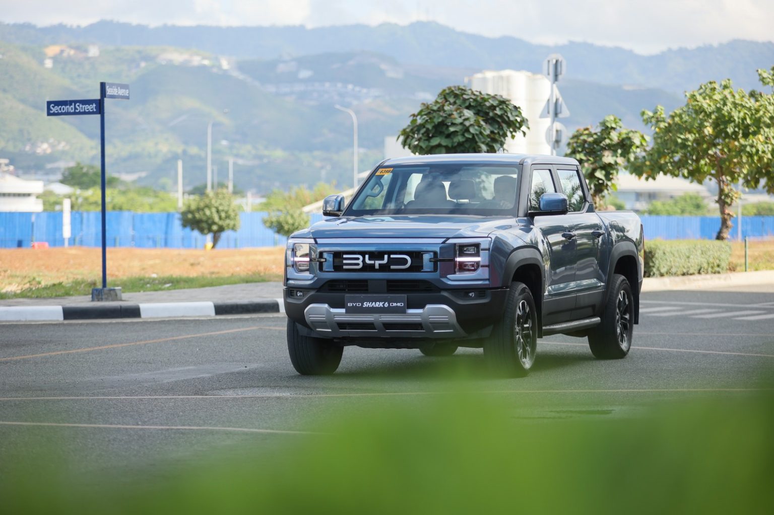 The BYD Shark 6 DMO is a plug-in hybrid electric pickup truck with Dual Mode Off-Road All Wheel Drive technology. The unit in this photo is seen being driven on a clean street, with out of focus greenery as foreground to give the photo a more creative touch
